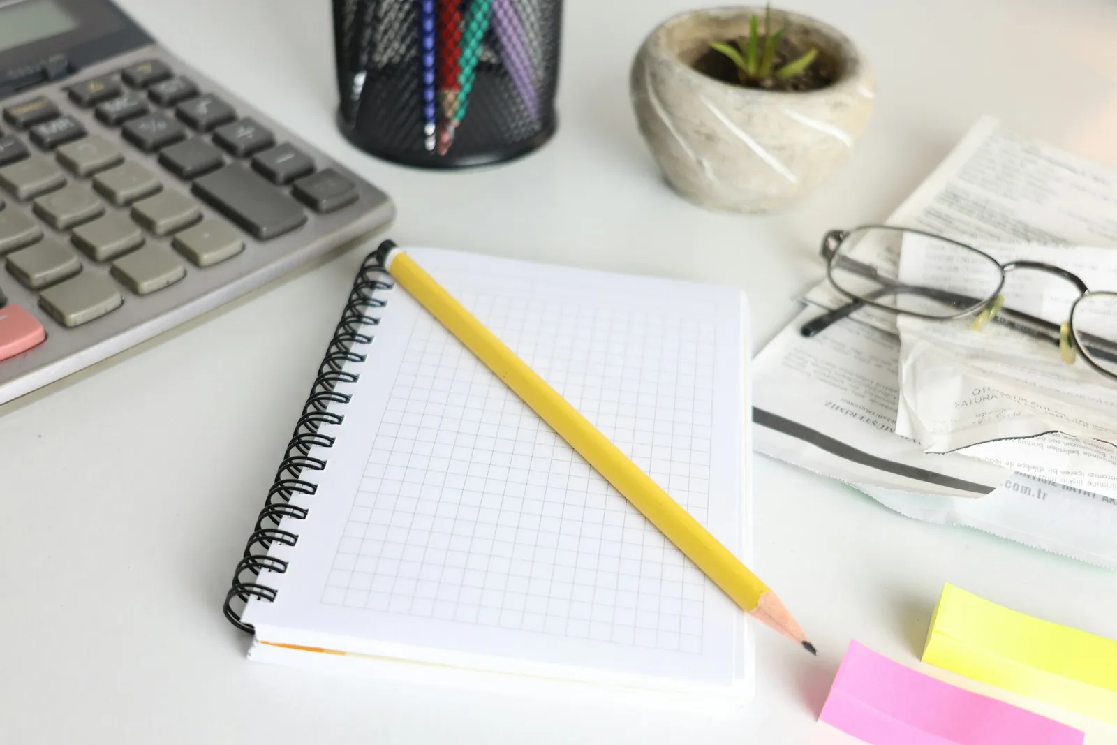 Overhead view of a desk with a calculator, an open notebook, a pencil, and eyeglasses.